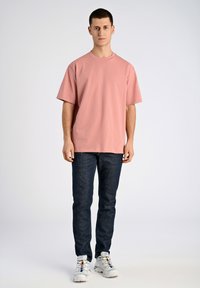 Ljusrosa bomullst-shirt med korta ärmar, rund hals, bärs tillsammans med mörkblå jeans och vita sneakers med färgglada detaljer.