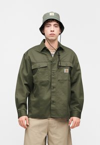 Πουκάμισο σε ελιά πράσινο με κουμπιά και δύο τσέπες στο στήθος, με εφαρμογή λογότυπου, σε συνδυασμό με καπέλο τύπου bucket στο ίδιο χρώμα, σε ουδέτερο φόντο.