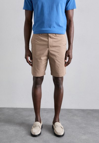 Beige Baumwollshorts mit flachem Bund, Seitentaschen und sichtbaren Nähten. Kombiniert mit einem blauen Rundhals-T-Shirt und beigen Slippern.