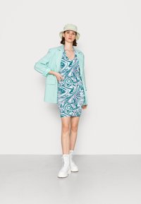 Blazer verde menta por cima de um vestido azul com padrão em espiral, combinado com botas brancas com atacadores e um chapéu de balde de cor clara. Texturas de tecido suaves evidentes.