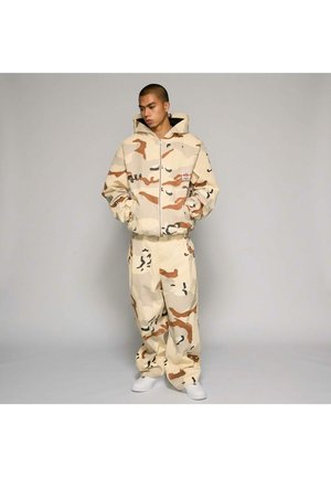 Mężczyzna stojący w beżowo-brązowej, pustynnej kamuflażowej kurtce z kapturem i pasujących szerokich spodniach oraz białych trampkach, z rękami w kieszeniach.
