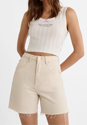 Beige denim shorts with frayed hems and front pockets, paired with a white ribbed crop top featuring a pink accent bow and embroidered text.
