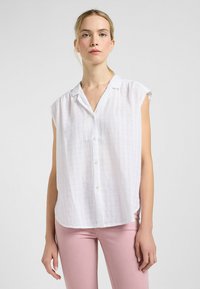 Blusa blanca de manga corta con cuello, que presenta una textura arrugada y cierre de botones, combinada con pantalones ajustados de color rosa claro.