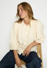 Light beige button-up shirt with puffed sleeves and a subtle leaf pattern, paired with dark, loose-fitting jeans.