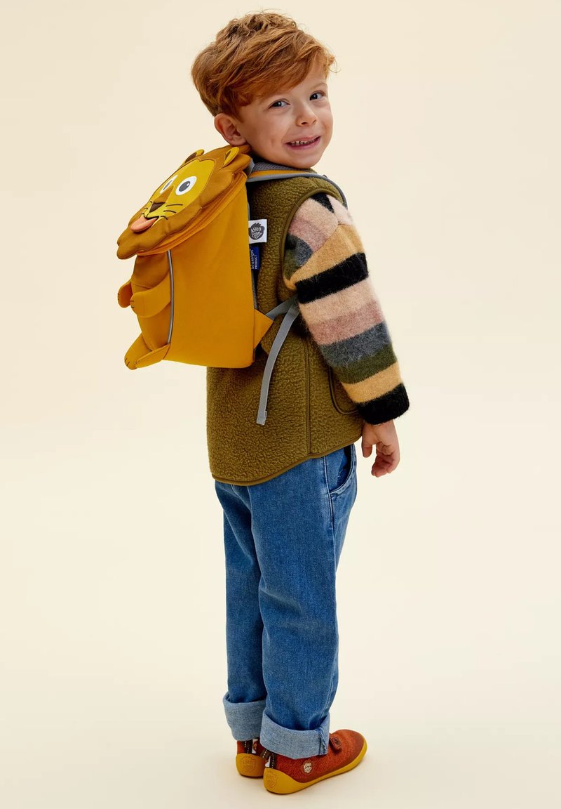 Young red-haired child wearing a striped sweater, green vest, rolled-up jeans, orange shoes, and a yellow lion-shaped backpack, smiling backward.