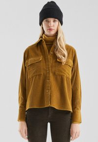 Senfgelbe Cordhemd mit zwei Brusttaschen, Knopfverschluss und langen �Ärmeln. Kombiniert mit einer schwarzen Strickmütze und einem Rollkragenpullover.