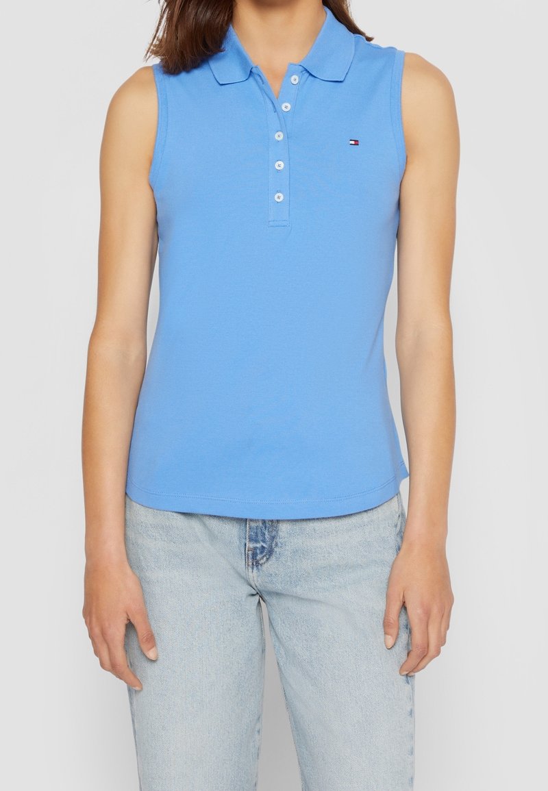 Polo sans manches bleu clair avec patte de boutons et petit logo sur la poitrine, porté avec un jean délavé clair.