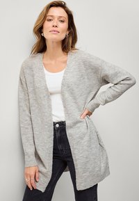 Cardigan lavorato a maglia grigio chiaro con apertura frontale, maniche a costine e vestibilità comoda, abbinato a una maglietta bianca e jeans scuri.
