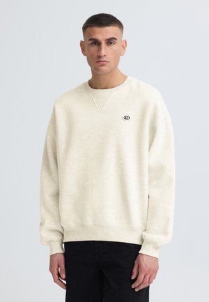 Man met kort haar die een crèmekleurige trui draagt met een klein "SD" logo en zwarte broek, staand tegen een effen lichte achtergrond.