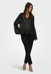 Camicia nera trasparente con pois, maniche lunghe e svolazzanti, orlo con bordi ondulati, abbinata a pantaloni neri e décolleté nudi.