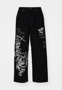 Jean large noir en denim, avec des impressions graphiques blanches comprenant du texte et des motifs le long des jambes. Comprend des poches avant et une fermeture à bouton.