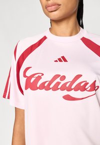 Rózsaszín sportos rövid ujjú póló piros díszítésekkel, amelyen az "adidas" logó piros színben jól láthatóan a mellkason szerepel.