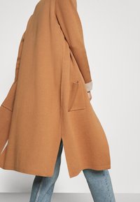 Abrigo largo de color marrón claro con un corte relajado, bolsillos laterales y un dobladillo redondeado, combinado con jeans azul claro. Confeccionado en una suave tela texturizada.