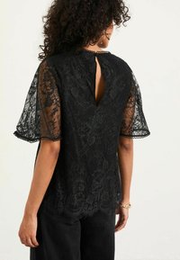 Schwarze Spitzenbluse mit Schlüssel-Ausschnitt im Rücken, ausgestellten transparenten Ärmeln, gewelltem Saum und floralen Mustern. Kombiniert mit dunklen Hosen.