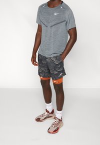 Man som bär en gråmönstrad Nike sporttröja, mörka shorts med spruckna mönster och orange kompressionsleggings, vita strumpor samt röda och beige löparskor.