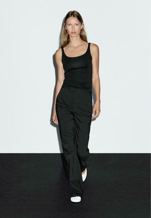 Femme marchant en avant portant un débardeur noir, un pantalon noir, des chaussures blanches et une bague en argent, devant un mur clair et un sol sombre.