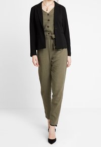 Blazer negro combinado con un mono verde oliva que cuenta con botones y un lazo en la cintura. La modelo lleva unos zapatos de tacón negro.