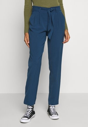 Persoon draagt blauwe broek met hoge taille en een gestrikte ceintuur, groene geribbelde lange mouwen top, en zwarte hoge sneakers met witte veters en zolen.