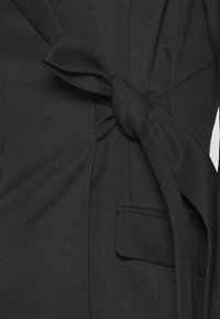 Blouse portefeuille noire avec un nœud à l'avant, en tissu lisse, coupe ajustée et détail de poche latérale.