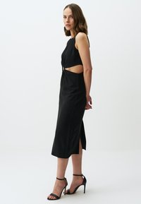 Robe noire sans manches avec des découpes sur les côtés et un détail torsadé à la taille. Longueur midi avec une fente sur le côté. Portée avec des escarpins noirs à talons aiguilles.
