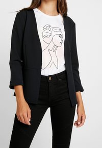 Blazer noir à manches trois-quarts porté sur un t-shirt blanc comportant un dessin minimaliste au trait d'un visage, assorti à un jean noir.