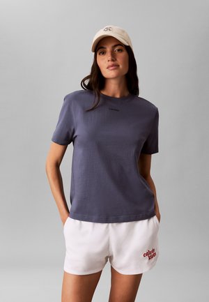 Frau mit beiger Kappe, dunkelgrauem Calvin Klein T-Shirt und weißen Calvin Klein Shorts, stehend mit Händen in den Taschen vor grauem Hintergrund.