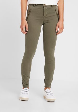 Frau trägt olivgrüne, schlank geschnittene Hose mit Reißverschlusstaschen vorne und weiße Turnschuhe, steht vor einem schlichten Hintergrund.