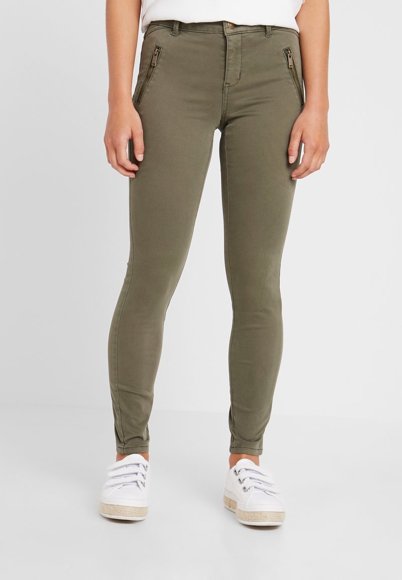 Femme portant un pantalon ajusté vert olive avec des poches avant zippées et des baskets blanches, debout devant un fond uni.