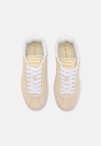 Sneakers Lacoste con tomaia in suede beige, lacci bianchi e suola in gomma. Presentano il logo sulla linguetta e una texture a motivo sul lato.