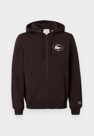 Man en vrouw staan naast elkaar, beiden dragen zwarte hoodies met rits, beige broeken, witte tops en sneakers; vrouw houdt een kleine zwarte handtas vast.