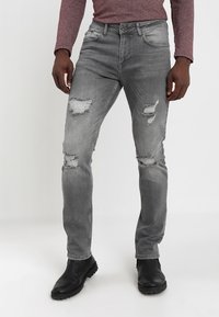 Jeans desgastados en gris con múltiples desgarros, un diseño ajustado y un acabado descolorido, combinados con botas de cuero negras. Detalles de costura sutiles.
