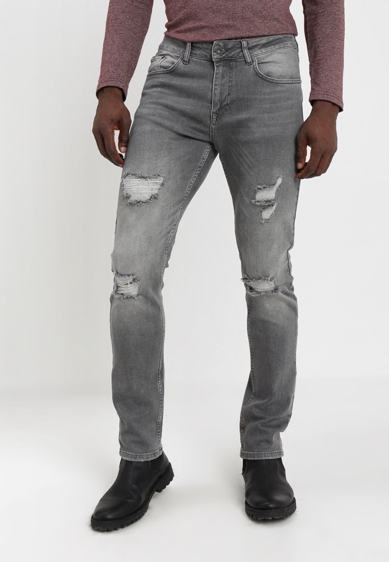 Jeans desgastados en gris con múltiples desgarros, un diseño ajustado y un acabado descolorido, combinados con botas de cuero negras. Detalles de costura sutiles.