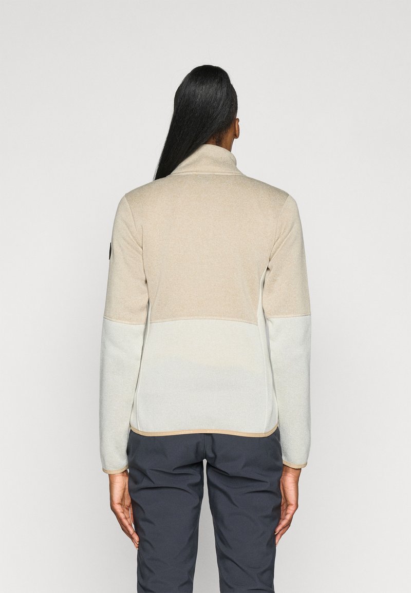 Chaqueta de forro polar beige y gris claro con cuello alto, bloqueos de color contrastantes y acentos cosidos. Combinada con pantalones gris oscuro.