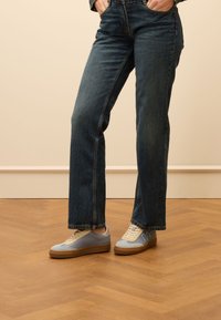 Blauwe en grijze sneakers met witte veters, een crèmekleurige zool en zijaccenten. Gecombineerd met donkerblauwe recht gesneden jeans op een houten vloer.