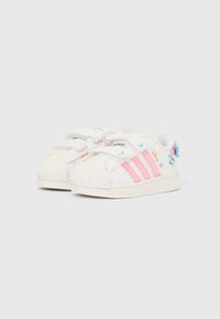 Biele tenisky pre batoľatá s ružovými pruhmi, modrým motýľom a potlačou postavičky, s dvoma suchými zipsmi na bielom pozadí.