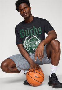 Sort grafisk t-shirt med grøn tekst "Milwaukee Bucks" og et cirkulært design, parret med grå shorts og sorte basketballsko. Holder en basketball.