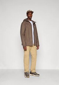 Barna vízálló dzseki kapucnival, fehér ing fölött viselve. A szettet bézs nadrág és sötét cipő fehér díszítésekkel egészíti ki.