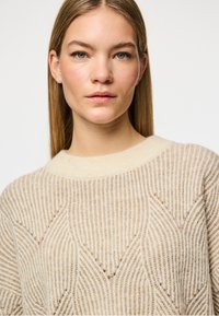 Beige strikket genser med ribbet tekstur og geometrisk mønster; har en bred rund hals og en myk finish.