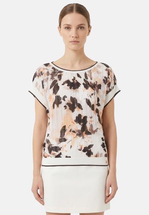 Donna che indossa un maglione a maniche corte con motivi floreali neri e pesca abbinato a una gonna bianca, in piedi contro uno sfondo semplice.