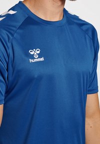 Modrá sportovní košile vyrobená z hladké látky, s kulatým výstřihem, krátkými rukávy a bílým logem a akcenty na ramenou.