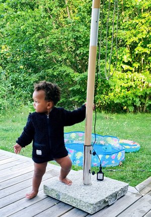 Lapsi mustassa fleece-takissa seisoo kivipohjalla ja pitää tikkua käsissään. Taustalla näkyy sininen puhallettava uima-allas.