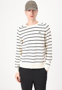 Pulôver listrado em creme e azul-marinho com punhos e bainha canelados, apresentando um pequeno logotipo verde da Lacoste. Usado com calças cinza-escuro e um boné.