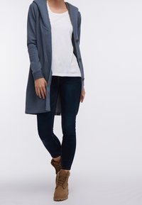 Person iført en lang blå cardigan over en hvid skjorte, mørke tætsiddende jeans og brune snørebåndssko, der står op ad en ensfarvet baggrund.