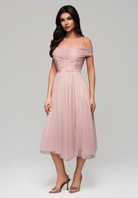 Robe rose décolletée avec un corsage froncé et un revêtement en tulle à pois, longueur mi-mollet, terminée par une ceinture en satin et des talons assortis.