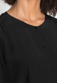 Blusa nera con bottoni, caratterizzata da un collo rotondo e dettagli arricciati sulle spalle, realizzata in tessuto leggero e testurizzato.