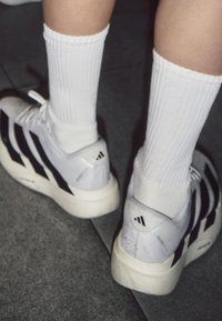 Personne portant des baskets blanches Adidas avec des bandes noires et des chaussettes blanches côtelées sur un sol gris.