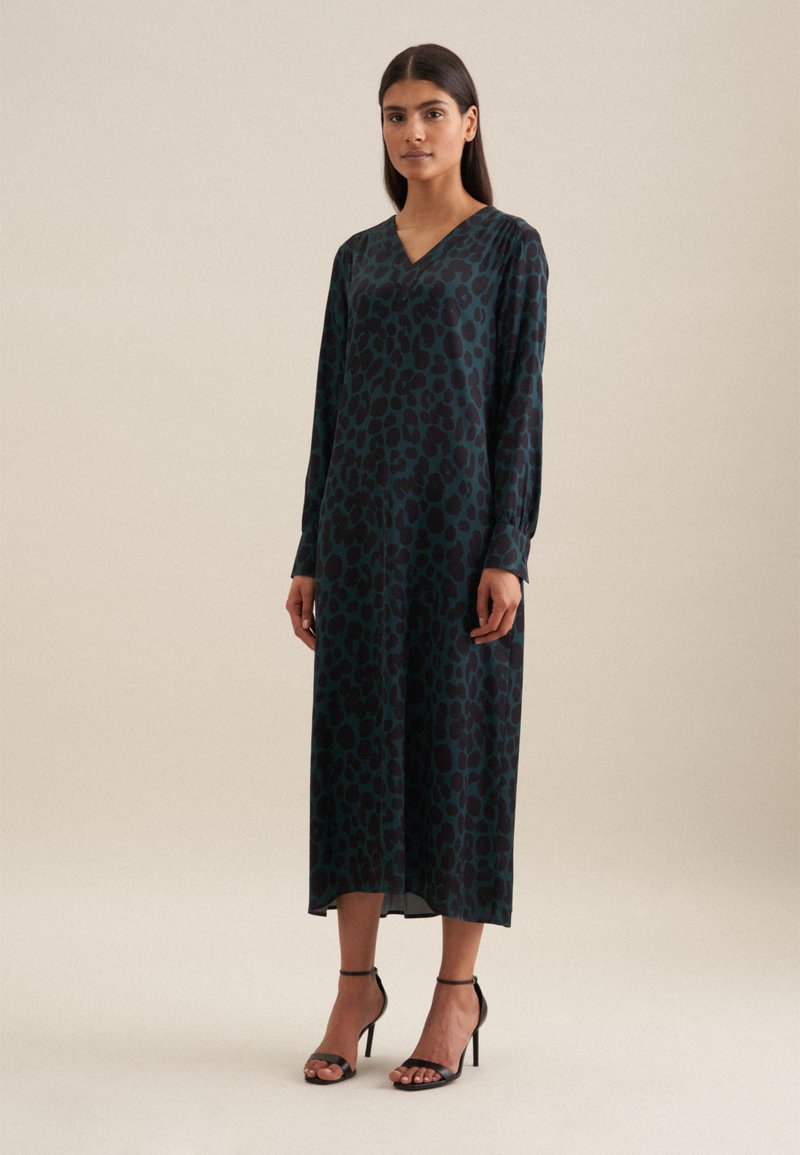 Langärmliges V-Ausschnitt-Kleid in Dunkelgrün mit schwarzen Leopardenflecken, ausgestattet mit einer fließenden Silhouette und geknöpften Ärmeln, kombiniert mit schwarzen High Heels.