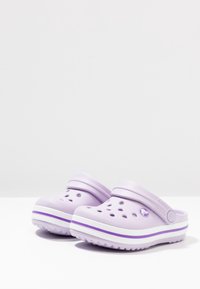 Lila crocs med ett perforerat mönster, mjukt material, en bakrem och vit-lila randig detalj längs sulan.