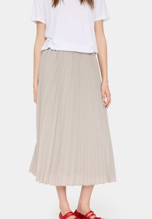 Vrouw draagt een beige geplooide midi-rok, een wit topje met korte mouwen en rode sandalen, staand tegen een effen lichte achtergrond.