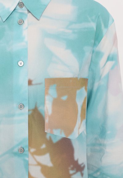 Lichtblauw shirt van een zachte, doorschijnende stof, met een beige borstzak en overlappende bladerenpatronen in subtiele tinten. Knopdetails zichtbaar.